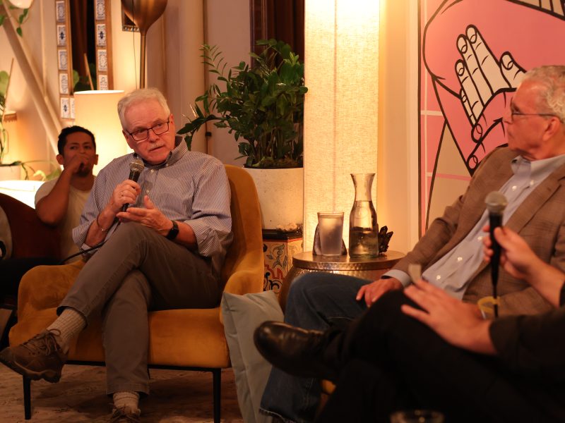 Two men are seated, speaking into microphones during a panel discussion in a cozy, well-lit room with plants and artwork in the background.