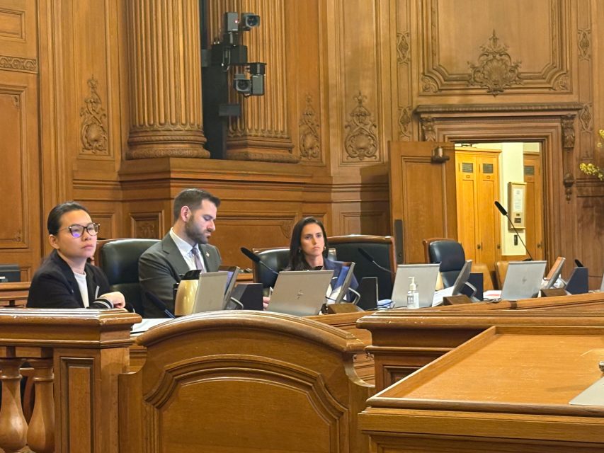 Three people in formal attire sit at a conference table with laptops and microphones in a wood-paneled meeting room.
