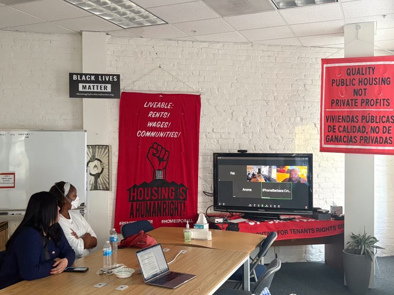 Two people sit at a table with laptops and water bottles, attending a Zoom meeting in a room decorated with banners about housing rights and social justice.