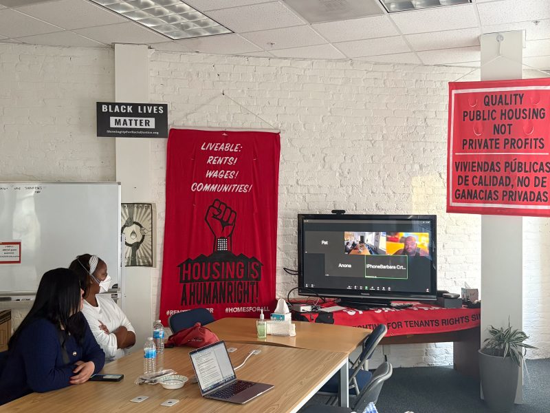 Two people sit at a table with laptops and water bottles, attending a Zoom meeting in a room decorated with banners about housing rights and social justice.