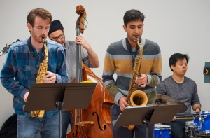 Four musicians perform indoors; two play saxophones, one plays an upright bass, and one plays drums. Sheet music is visible on stands in front of the saxophonists.