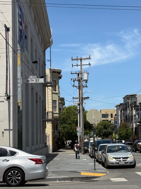 A city street intersection with cars parked along the side, a person walking on the sidewalk, and a street sign reading "16th St" under a clear blue sky.