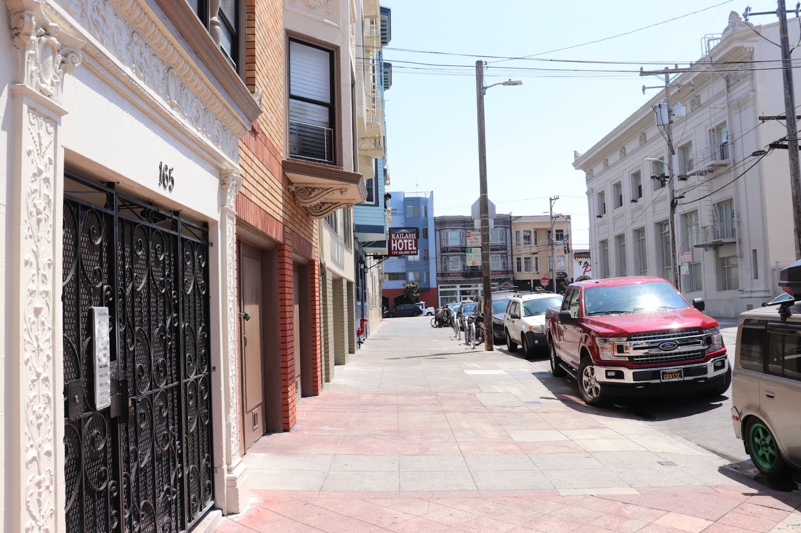 A city sidewalk with parked cars, buildings lining both sides, and a red hotel sign visible in the distance under a clear sky.