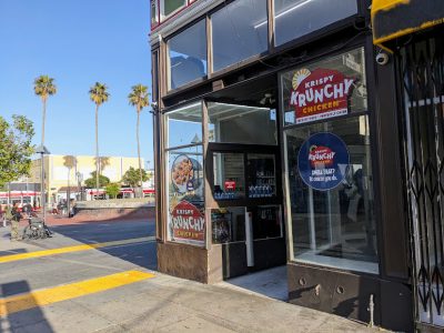 Street view of a Krispy Krunchy Chicken storefront with signs on the windows, located at a corner in a sunny urban area with palm trees and people nearby.