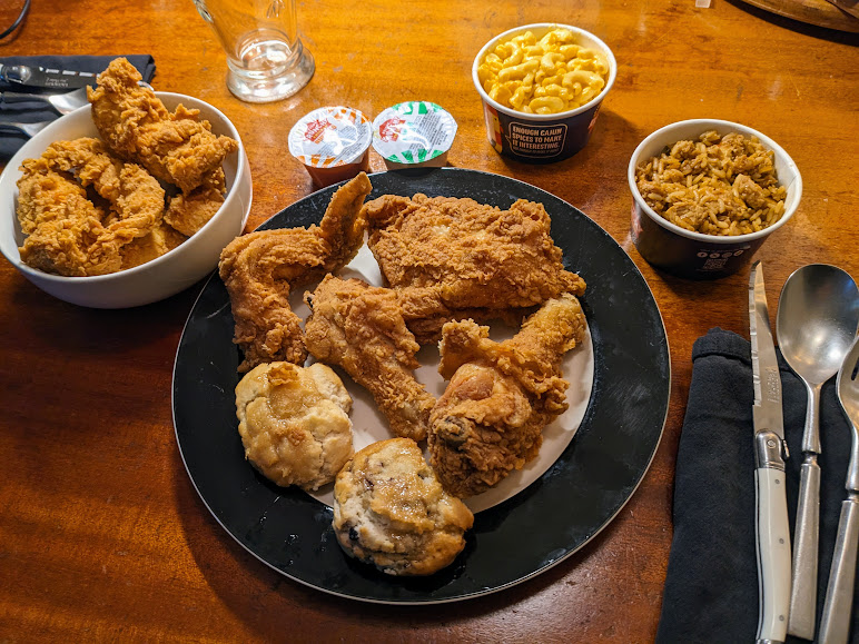 A plate of fried chicken, two biscuits, a bowl of fried chicken pieces, a cup of macaroni and cheese, a cup of rice, sauce containers, and cutlery on a wooden table.