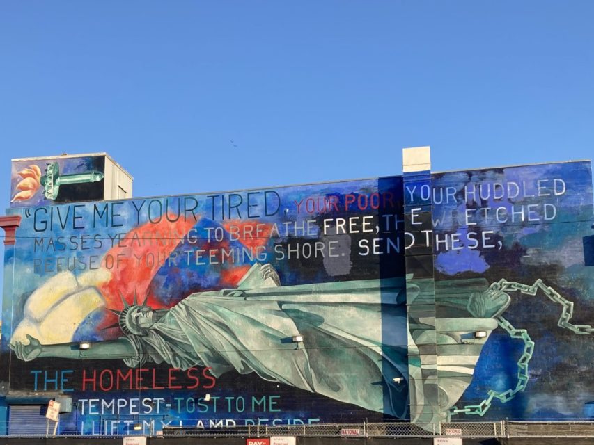 A mural on a building depicts the Statue of Liberty with broken chains, featuring the poem "The New Colossus" and bolded words: "the homeless," "your poor," and "free.