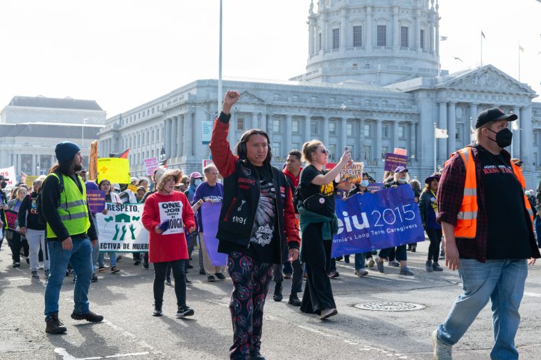 A diverse group of protesters march outside a government building holding signs and banners, with one person raising a fist in the air.