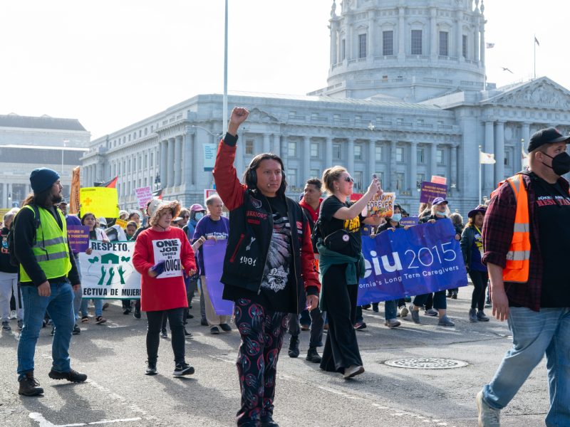 A diverse group of protesters march outside a government building holding signs and banners, with one person raising a fist in the air.