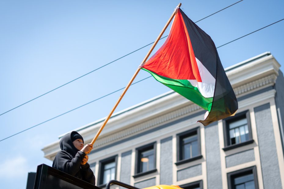 A person wearing a black hoodie stands on a vehicle and holds up a large Palestinian flag in front of a gray building.