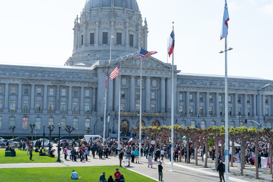 A crowd gathers outside a large government building with columns, domed roof, and multiple flags, for a May Day demonstration.