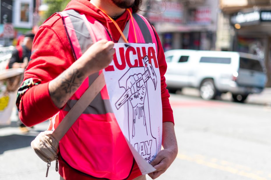 A person in a red hoodie and reflective vest holds a protest sign with an illustration of a raised fist holding a pen and partially visible text reading "LUCHA.