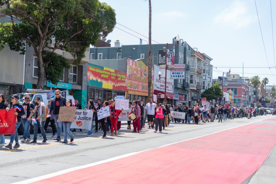 A group of people march down a city street holding signs and banners during a protest or demonstration on a sunny day.