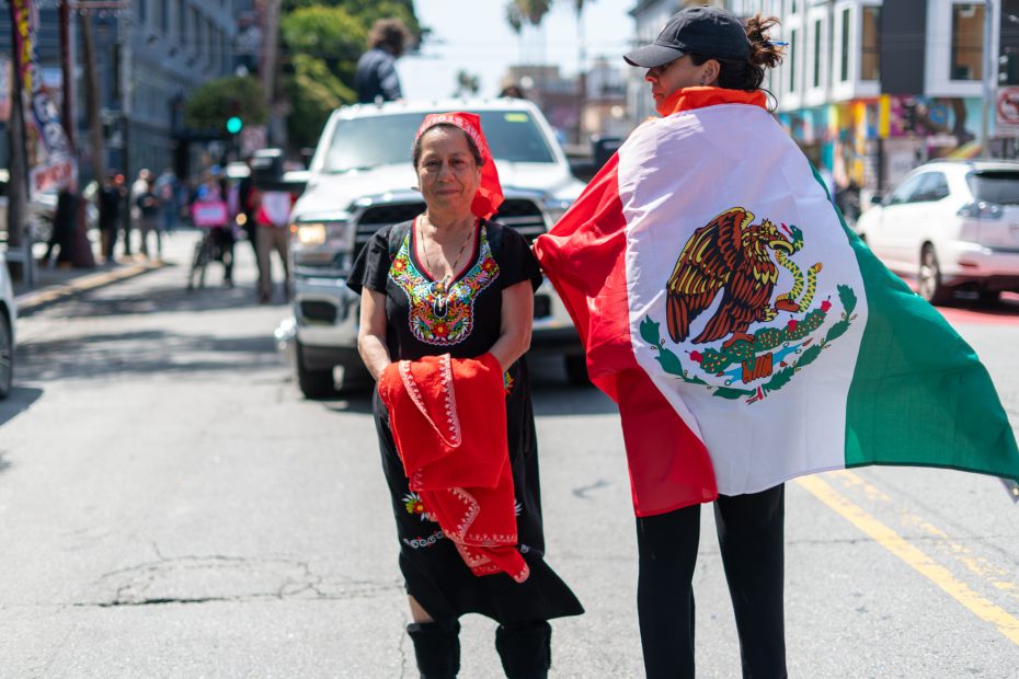 Two people stand on a city street; one wears traditional clothing and holds a red cloth, while the other is draped in a Mexican flag. A vehicle and other people appear in the background.