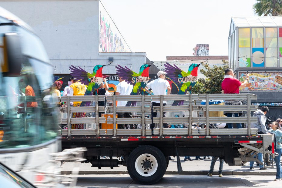 A parade float with colorful hummingbird wings and people standing on it moves down a city street; a bus passes in the foreground.