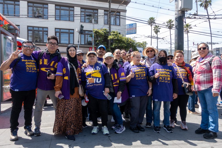A group of people wearing matching purple SEIU shirts pose together on a city sidewalk under a sunny sky.