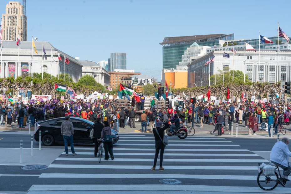 A large crowd gathers at a city plaza for a protest; people hold signs and flags while others observe or cross the street in the foreground.