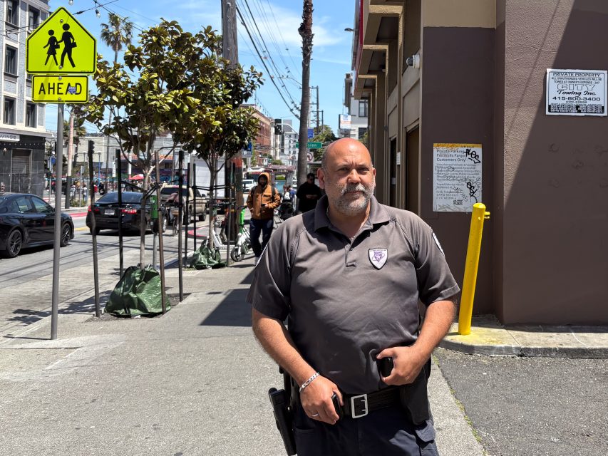 A man in a security uniform stands on a city sidewalk near a crosswalk sign on a sunny day.