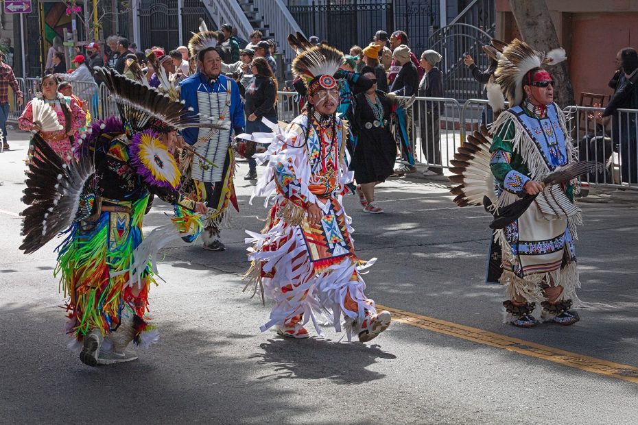 Three people in colorful traditional Native American regalia participate in a street parade, with onlookers and fences in the background.