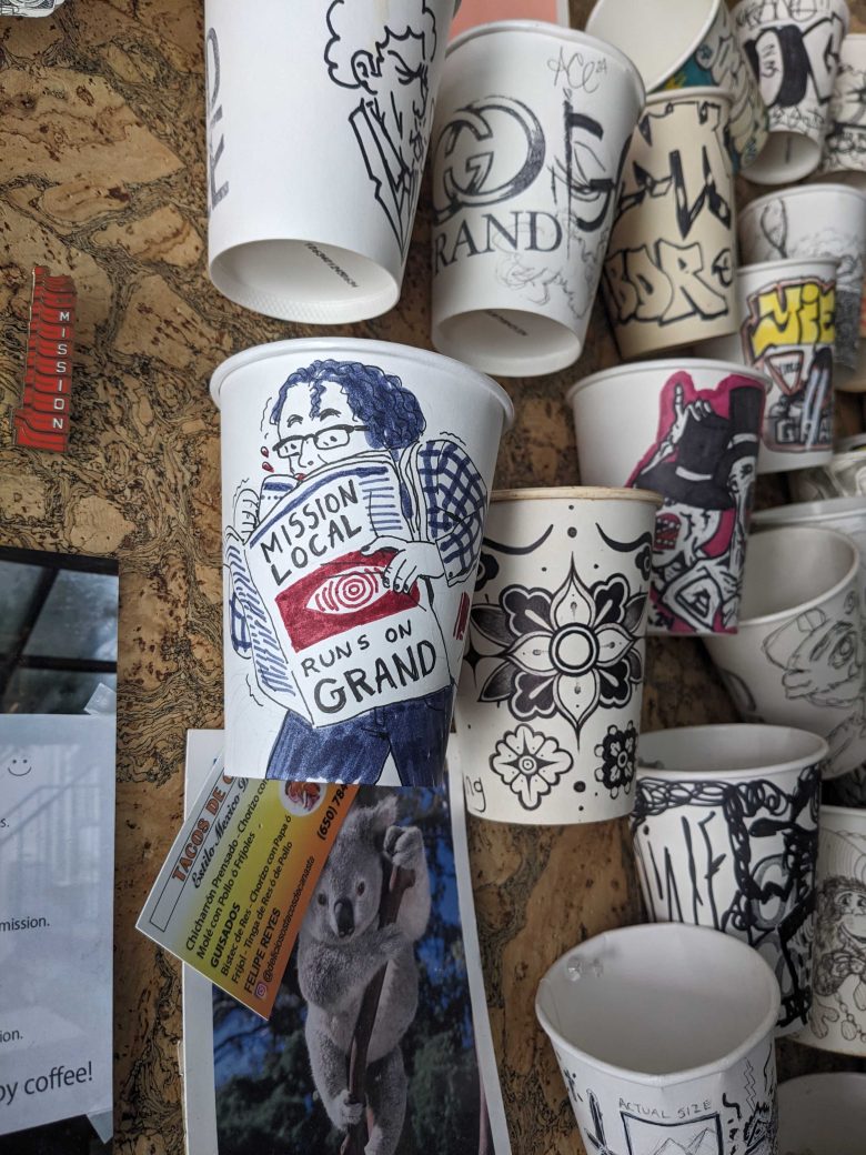 A display of decorated paper cups, featuring one cup with a hand-drawn person holding a newspaper titled "Mission Local Runs on Grand," among other cups with various designs.