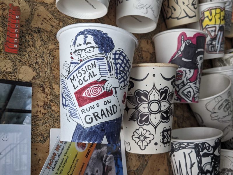 A display of decorated paper cups, featuring one cup with a hand-drawn person holding a newspaper titled "Mission Local Runs on Grand," among other cups with various designs.