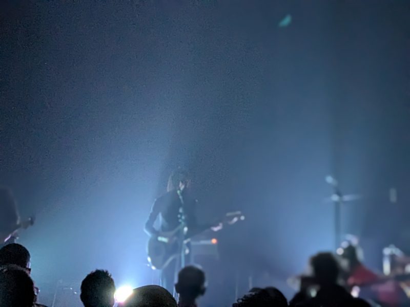 A musician plays guitar on a dimly lit stage with bright backlighting, while silhouettes of audience members are visible in the foreground.
