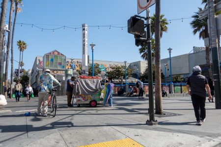 People walk and stand near a colorful street cart in a busy urban plaza with murals, palm trees, and bright sunlight.