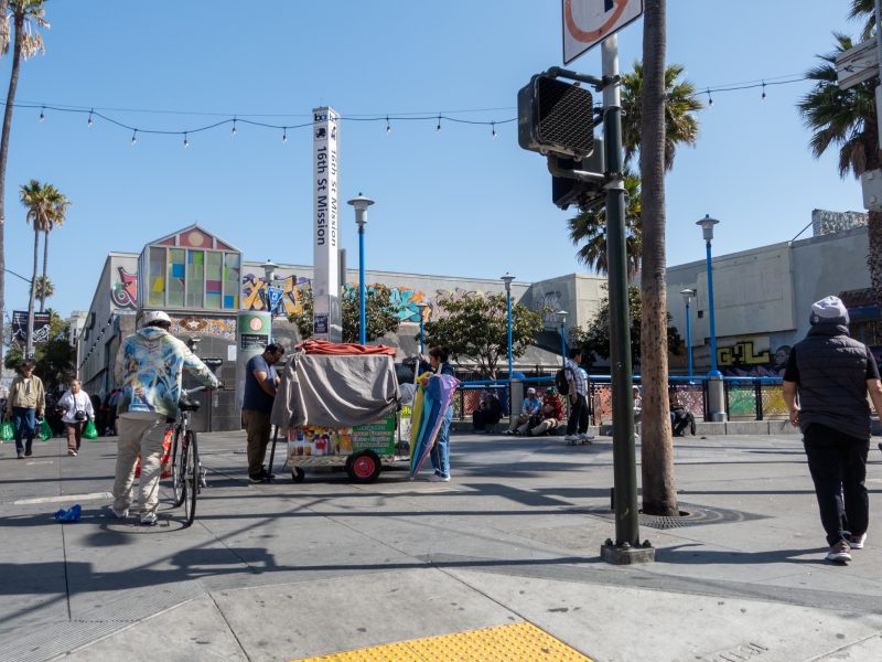 People walk and stand near a colorful street cart in a busy urban plaza with murals, palm trees, and bright sunlight.