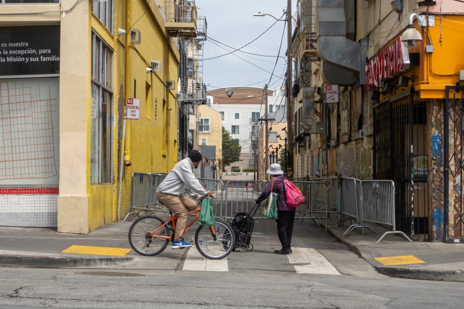 A man on a bicycle and a woman with a walking cart cross a street at a crosswalk between two buildings on a cloudy day.