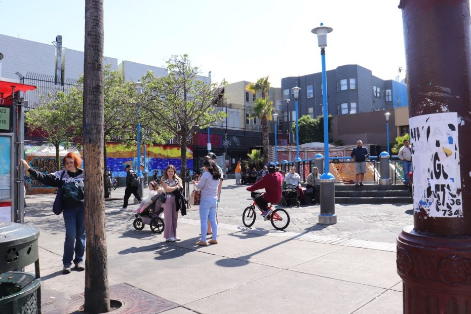 People walking and biking in a city plaza with colorful murals on the walls and a mix of sun and shade.