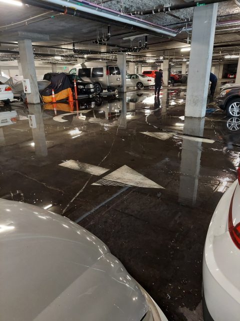 Flooded underground parking garage with cars parked, water pooled on the floor, a person standing, and large white arrow painted on the ground.
