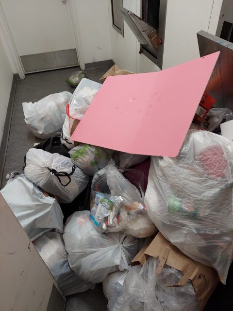A pile of trash bags, paper bags, and a large pink cardboard sheet are stacked in a hallway near a closed door and a trash chute.
