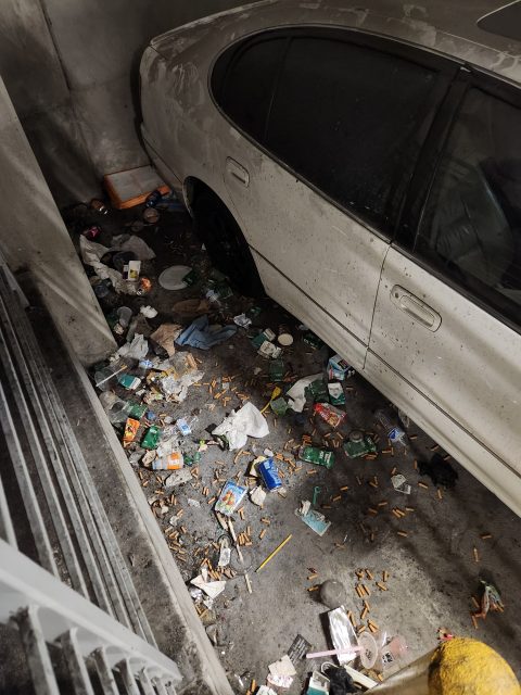 A dirty white car parked in a concrete garage next to a large pile of litter, cigarette butts, and trash scattered on the ground.