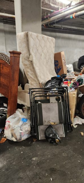 A pile of discarded items in a parking garage, including a mattress, folding chairs, wooden furniture, trash bags, and cardboard boxes.