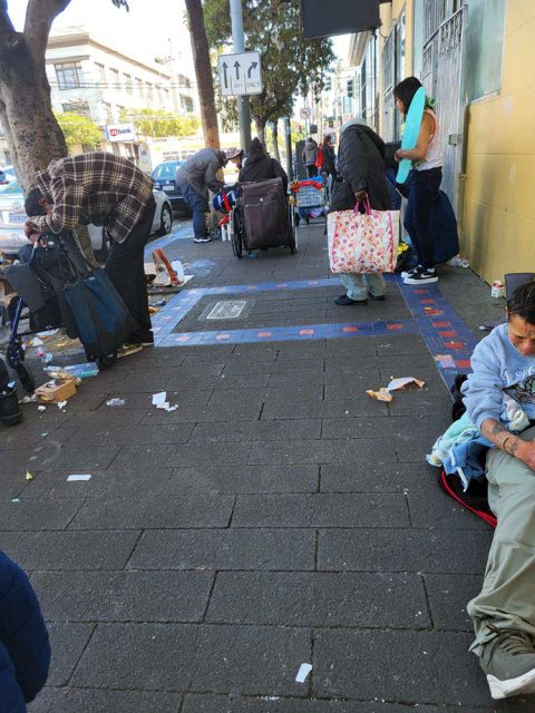 People with bags and personal belongings gather on a city sidewalk scattered with litter, some sitting while others stand or sort through items near a building.