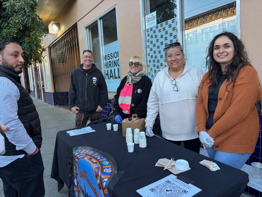 Five people stand around a table with paper cups and a coffee container on a city sidewalk near a building with “Mission Hiring Hall” signage in the window.
