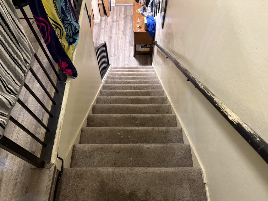 View looking down a carpeted staircase with a handrail on the right, leading to a wooden floor and desk at the bottom. Towels or clothes hang on the left railing.
