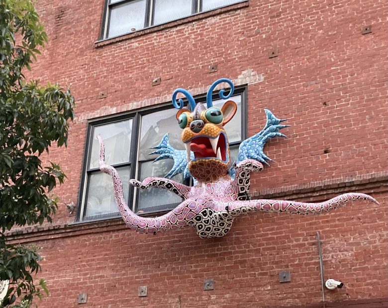 A colorful, whimsical creature sculpture with tentacles and large eyes is mounted on a brick building below a window.