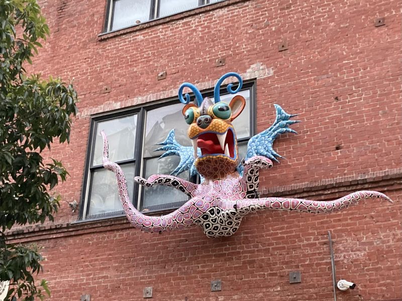A colorful, whimsical creature sculpture with tentacles and large eyes is mounted on a brick building below a window.