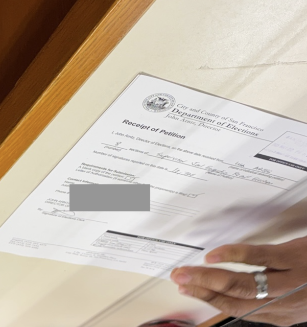 A person's hand holds a petition receipt form for the recall Engardio effort from the San Francisco Department of Elections on a table, with certain details redacted for privacy.