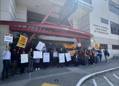 A group of people stand outside John O'Connell High School of Technology holding protest signs about education and school safety.