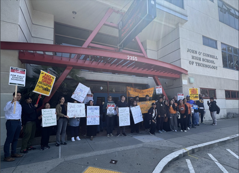 A group of people stand outside John O'Connell High School of Technology holding protest signs about education and school safety.