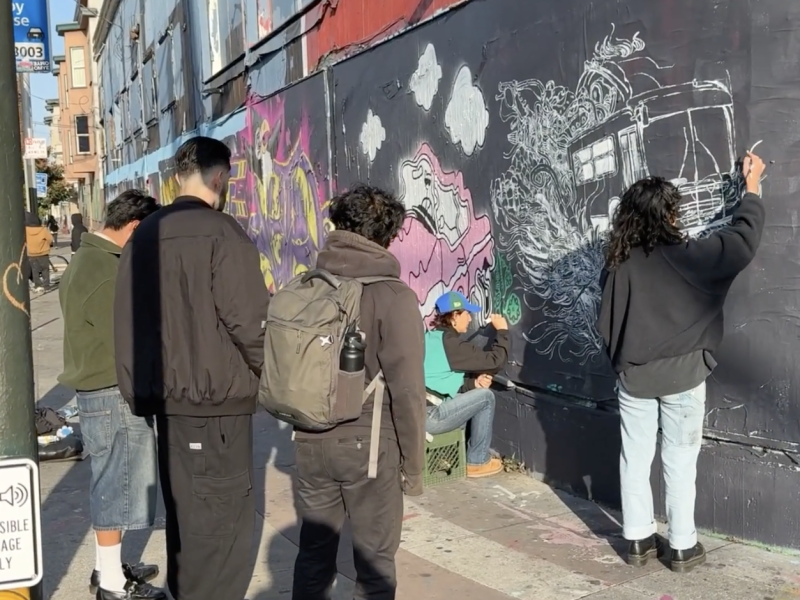 Several people stand and observe as a person draws intricate chalk designs on a black wall outdoors. One person sits nearby. The scene takes place on a sunny city sidewalk.