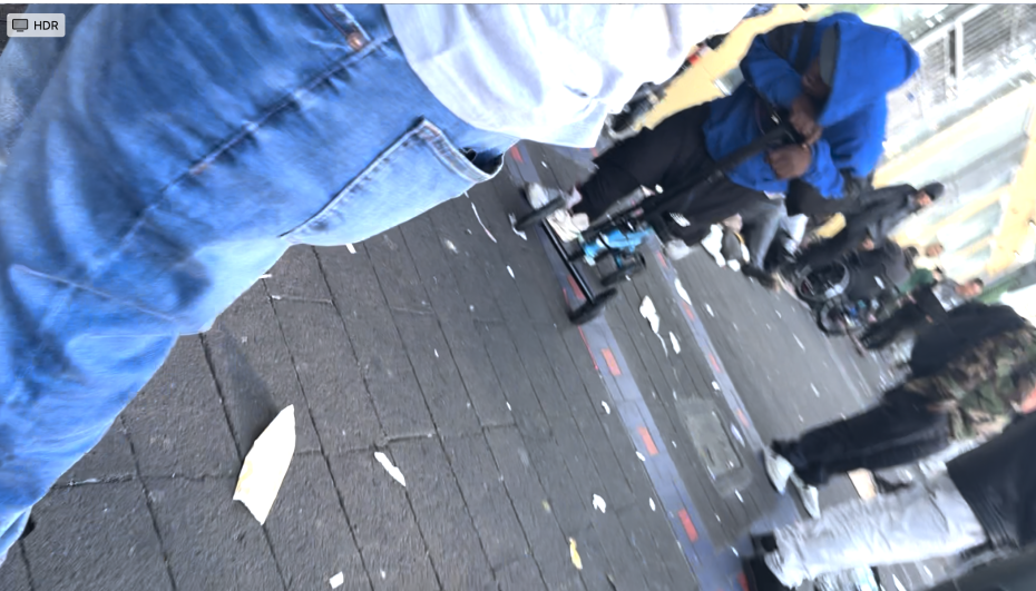 A busy urban sidewalk with several people standing and scattered litter on the ground. One person in a blue hoodie is leaning over a bicycle.