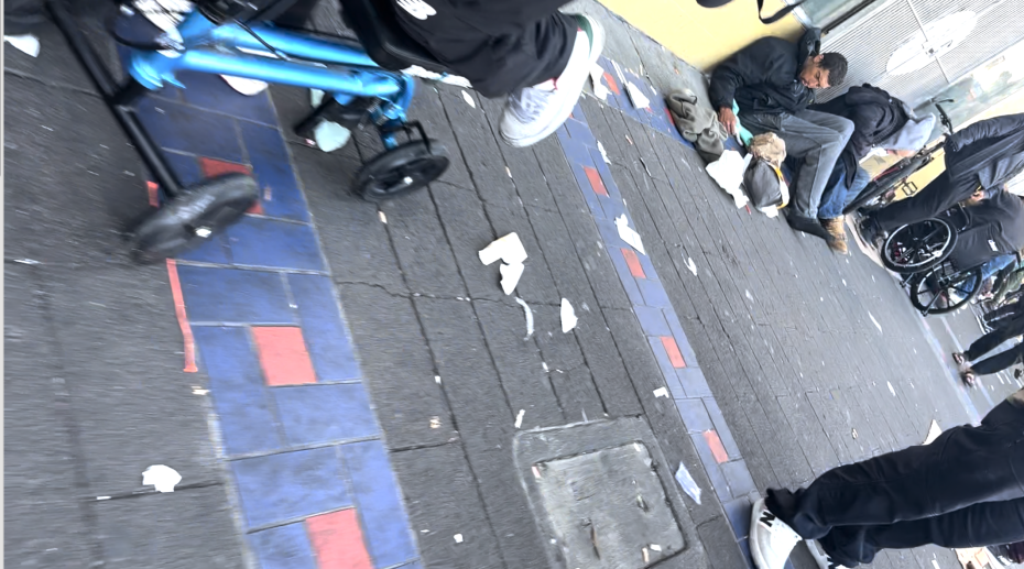 People are sitting and lying on a littered sidewalk next to a building; part of a wheelchair and stroller are visible in the foreground.