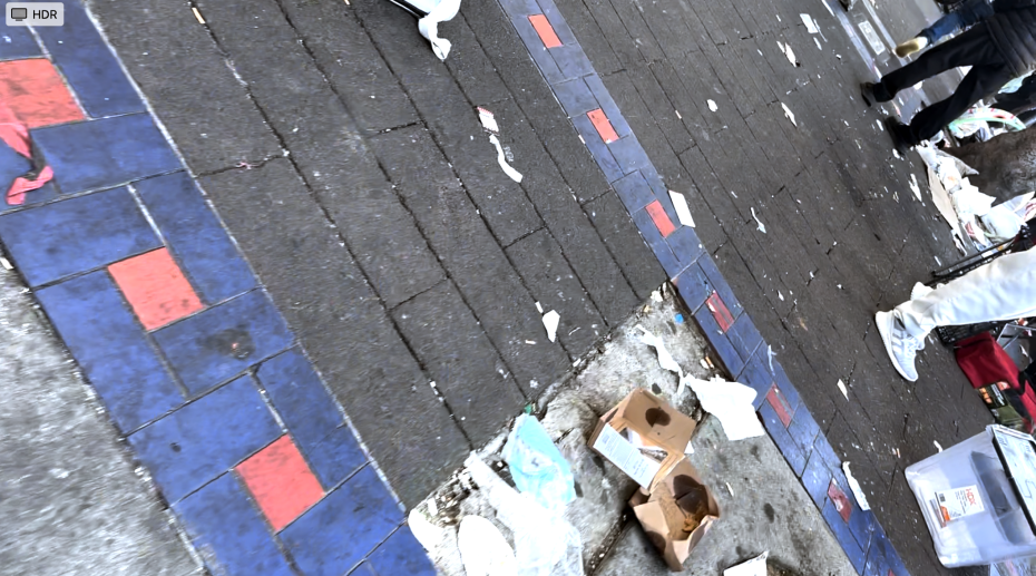 Litter, cardboard boxes, and plastic packaging scattered on a city sidewalk paved with blue and red tiles; several people’s legs visible in the frame.