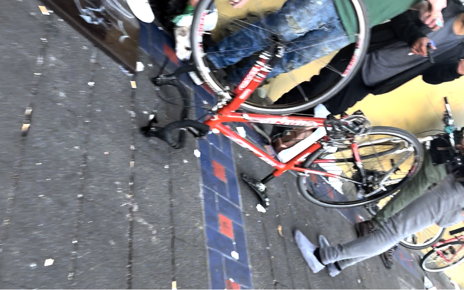 A red road bicycle with missing wheels lies on the sidewalk near two people standing, one barefoot, and one holding a phone.