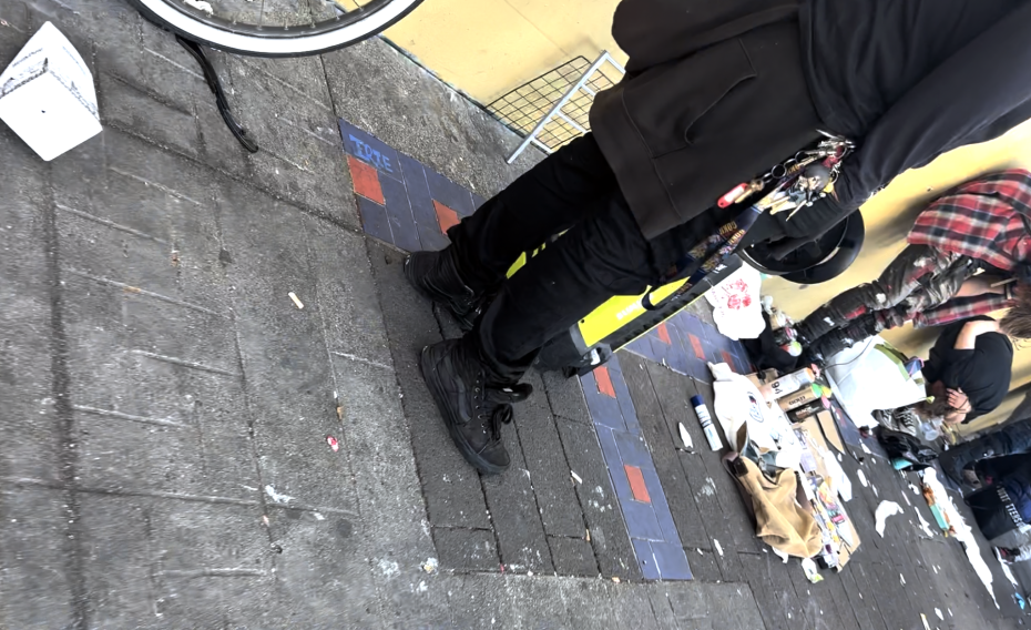People standing and sitting on a littered city sidewalk beside a yellow wall; various items, bags, and scattered trash are visible on the ground.
