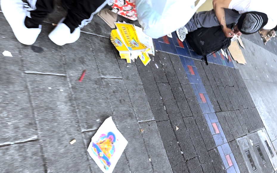 Two people sit and stand on a sidewalk with scattered items, including coloring books, paper, and bags, next to tiled pavement.