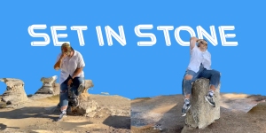 Two people sit on large rock formations under a clear blue sky with the text "SET IN STONE" in bold white letters above them.