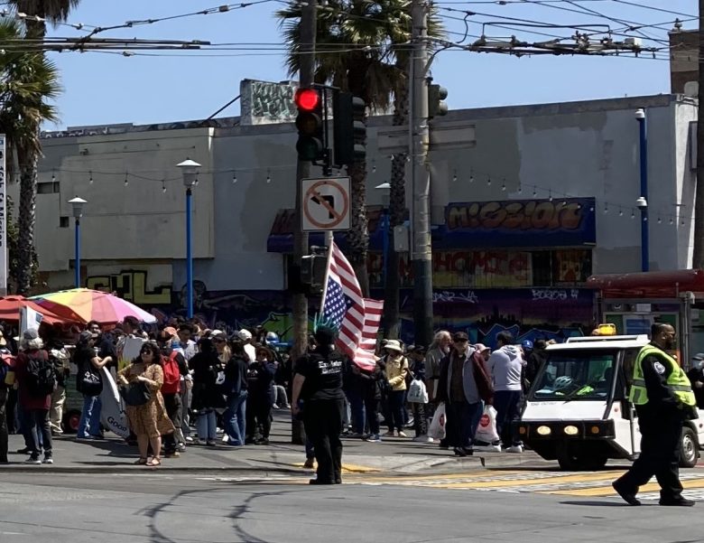 A crowd gathers at a street corner near a police officer, a small vehicle, and a person holding an American flag under sunny skies. Urban buildings and palm trees are in the background.